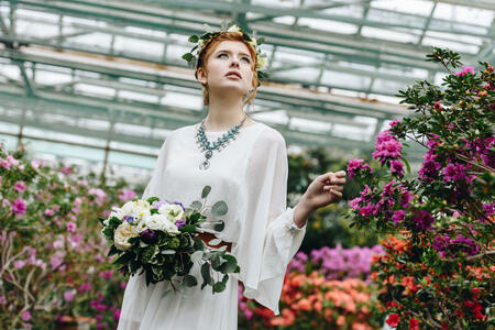 beautiful young woman in white dress and floral wreath holding wedding bouquet in botanical garden