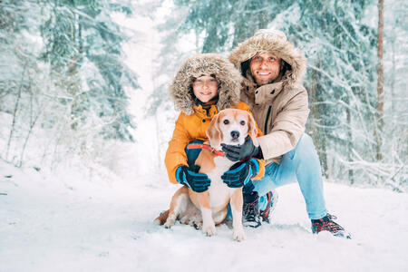 Father and son  dressed in Warm Hooded Casual Parka Jacket Outerwear walking with their beagle dog in snowy forest cheerful smiling faces portrait. Pets in family and winter outfit concept image.