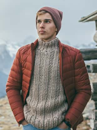 Handsome male model wearing warm sweater, winter coat and hat posing over mountains with snow. Active fashion clothes for cold weather.