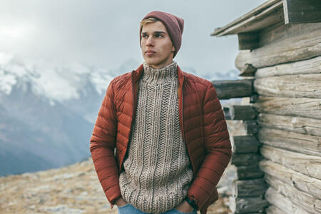Handsome male model wearing warm sweater, winter coat and hat posing over mountains with snow. Active fashion clothes for cold weather.