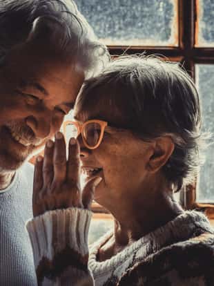 Senior old couple in love at home with snow winter outside the window. Aged woman whisper at man ear. Mature couple enjoy relationship and holidays indoor. Man and woman elderly smile