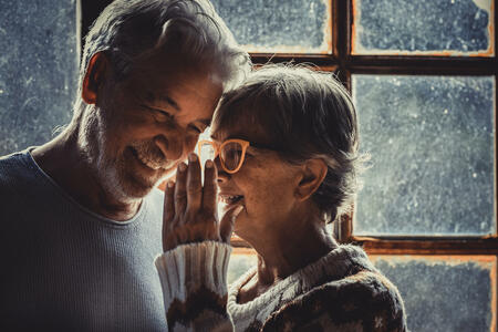 Senior old couple in love at home with snow winter outside the window. Aged woman whisper at man ear. Mature couple enjoy relationship and holidays indoor. Man and woman elderly smile