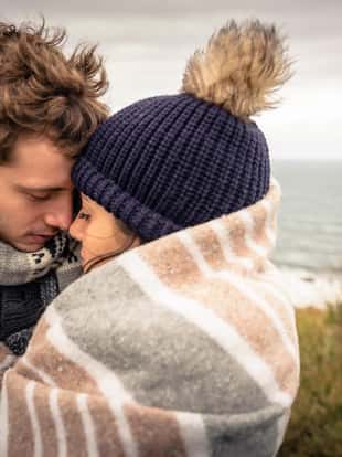 Closeup of young beautiful couple embracing under blanket in a cold day with sea and dark cloudy sky on the background