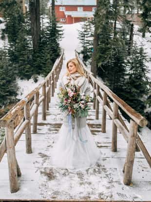 Beautiful woman stands on the wooden bridge in snowy forest in grey wedding dress with bouquet in hands