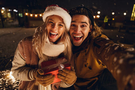 Cheerful young couple dressed in winter clothing holding gift boxes sitting outdoors, taking a selfie, snowfall