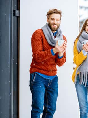 Portrait of a young lovely couple dressed in colorful sweaters standing with hot drinks on the terrace of the modern house at the mountains