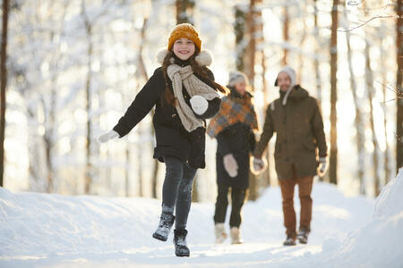 Full length portrait of happy little girl running towards camera in winter forest while enjoying walk with family, copy space