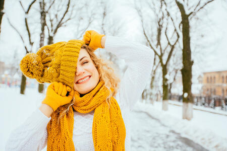 Outdoor close up portrait of blond curly playful hipster woman on street, looking at camera, smiling at snowy park. Model wearing white sweater, yelow winter hat, sarf, gloves. City lifestyle