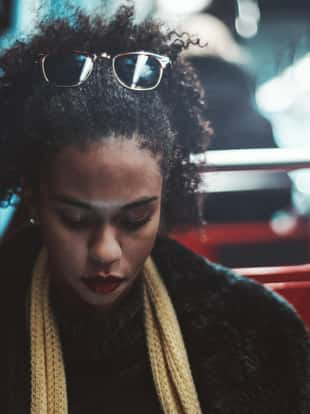 A pretty young African-American woman in a subway train is pensively looking down while working with her gadget; cute curly-hair Brazilian female in a demi-season coat on a seat of a metro carriage