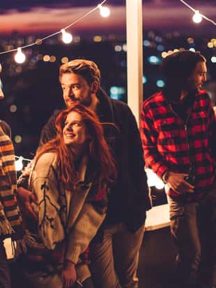 Couple of friends having a party on the rooftop of the building at sunset. Drinking bottles of beer and taking. Wearing knitted sweaters, hats and scarfs. They are happy and joyful.