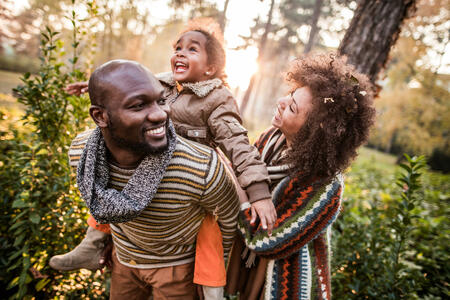 Happy African American family having fun in autumn day at the park. Father is piggybacking little girl.