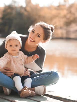Smiling woman holding her baby girl 1 year old sitting on wooden pier in park wearing autumn casual clothes. Looking at camera. Motherhood.
