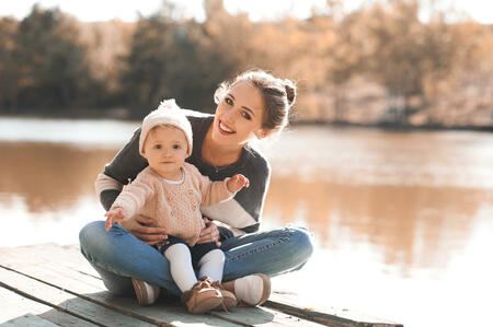 Smiling woman holding her baby girl 1 year old sitting on wooden pier in park wearing autumn casual clothes. Looking at camera. Motherhood.