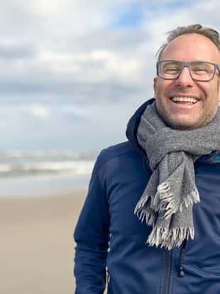 Happy laughing middle-aged man wearing glasses and a knitted woollen scarf standing on a deserted autumn beach on a cloudy day with copy space