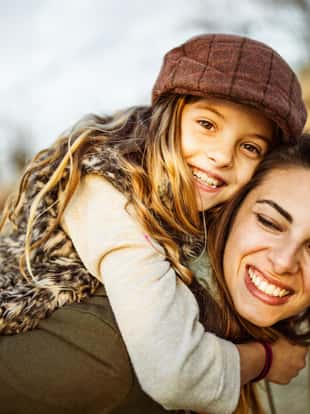 Young happy mother piggybacking her daughter in autumn day outdoors. Girl is looking at camera.