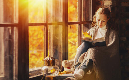 Happy young woman reading a book by the window in the fall