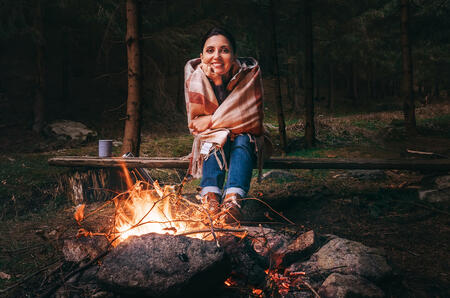 Pretty smiling young woman sits near the campfire in twilight autumn forest