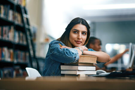 Shot of a young woman resting on a pile of books in a college library and looking thoughtful