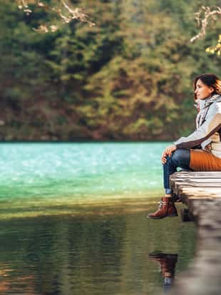 Woman traveler sits on wood bridge and looks calm on mountain lake at sunny autumn day