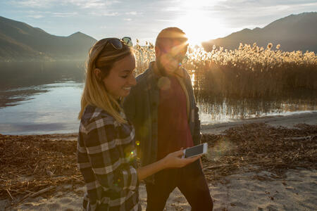 Cheerful young couple text messaging on smart phone by the lakeshore at sunset, beautiful Autumn sunlight.