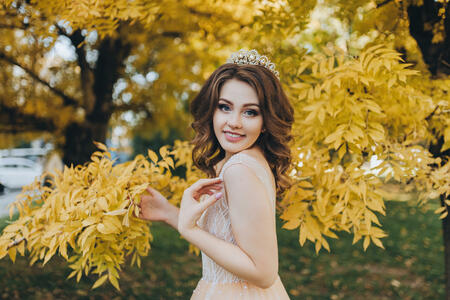 A smiling bride in a beige dress is standing in a park with yellow leaves. The princess with a crown enjoys the fall. Wedding portrait of a beautiful bride with brown hair. Wedding photography.