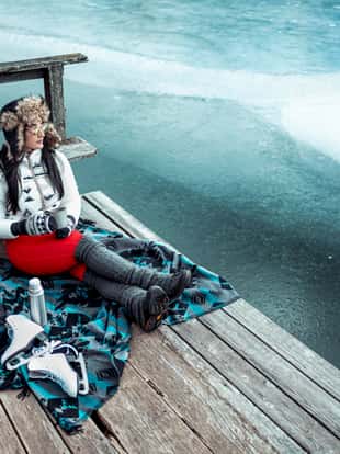 A woman sitting on a blanket on a pier next to the frozen lake. She is drinking a hot beverage.