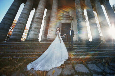 Panoramic view of a fairytale newlywed couple holding hands in front of old baroque ghotic church with columns at sunset