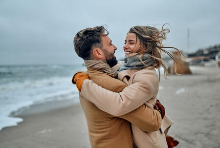 Beautiful married couple cuddling on the seashore, wearing a coat in the cold season.