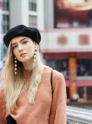 A beautiful young blonde woman dressed in a large warm pink sweater with a black beret and gorgeous earrings. She goes through a big spectacular Chinese city in autumn and enjoying every moment.