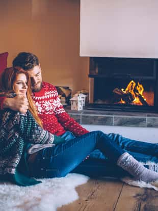 Couple on vacation at mountain cabin. Sitting on the floor on a fur by fireplace in cozy living room on Christmas. Wearing festive knitted sweaters and socks. Austrian Alps.