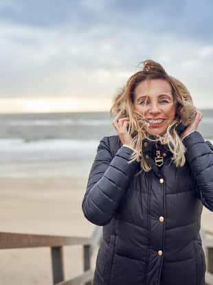 Middle-aged woman braving a cold winter day at the seaside standing on a wooden deck overlooking the beach on a breezy day smiling happily at the camera