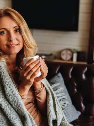 Middle aged woman relaxing at home with cup of coffee while sitting on sofa and covers with blanket. Looking in camera
