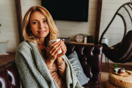 Middle aged woman relaxing at home with cup of coffee while sitting on sofa and covers with blanket. Looking in camera