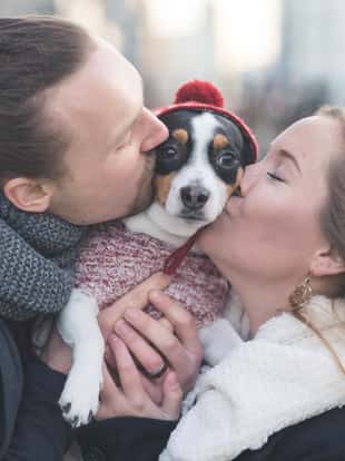 A young hipster couple poses with their dog for an adorable portrait in a downtown shopping district. Their pet has a warm Christmas hat on. They are each kissing his cheek as he looks with a serious expression at the camera.
