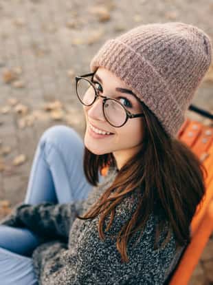 Rear view image of beautiful young woman smiling broadly, wearing eyeglasses, knitted sweater and hat. Outdoor portrait of pretty female model sitting on the bench in the street.
