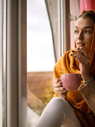 Copy space shot of smiling young woman wrapped in a cozy yellow scars, relaxing and enjoying a warm cup of tea while looking through the window and reminiscing on old memories.