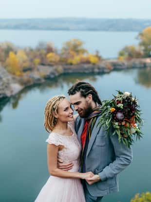 Stylish couple newlyweds smile and hugging standing before a lake. Bride and groom with dreadlocks look at each other. Autumn wedding ceremony outdoors. Close-up portrait
