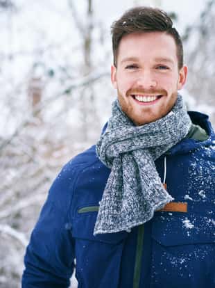Young bearded man in warm coat smiling at camera on blurred background of snowy forest.
