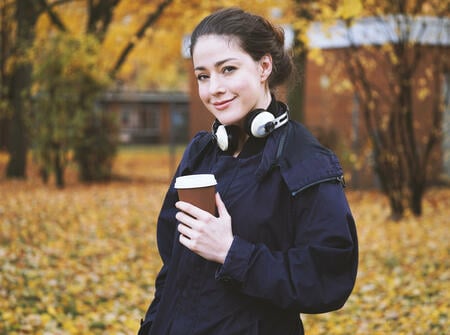 trendy young woman with headphones around her neck and coffee to go cup in her hand walking in park on a cold autumn day