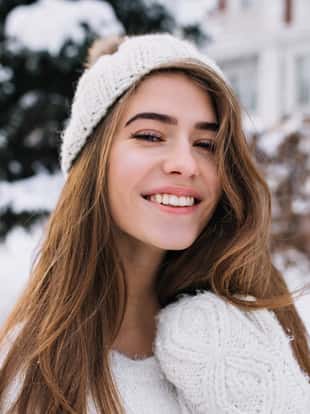 Close-up portrait of pleased blonde woman with sincere smile enjoying winter morning. Lovely european girl in white hat looking at snowy view outdoor..
