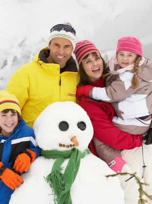 Family Building Snowman On Ski Holiday In Mountains Smiling To Camera