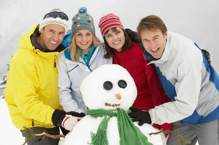 Group Of Friends Building Snowman On Ski Holiday In Mountains Having Fun And Smiling To Camera