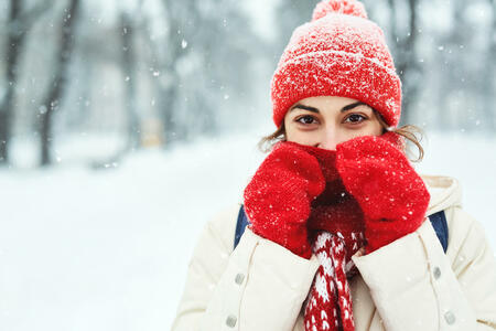 closeup portrait of a smiling woman in warm clothes, red knitted cap, scarf and mittens walking on the snowy street under falling snowflakes after blizzard in city. Happy woman playing with the snow