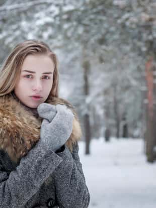 A stylishly dressed, beautiful girl in a gray coat with a fur collar stands alone in a winter forest, warming her hands in fur mittens. Snowflakes fall on her shoulders.