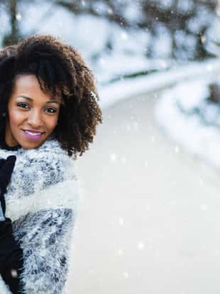 Cheerful stylish afro hair woman portrait under the snow at winter mountain road. Female wearing warm clothes and gloves.