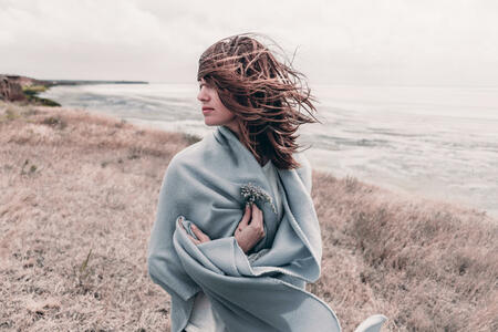 Attractive young woman standing on a windy cold beach wrapped in warm blanket with flower in her hand. Windy cold weather concept.