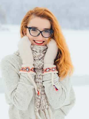 Beautiful smiling red head woman with glasses is wrapped in the scarf during the snowfall in the forest. Half-length horizontal portrait