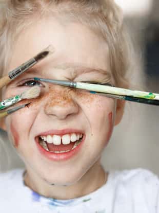 Blonde little girl in playful mood with paint on her freckled face and blue eyes covering her face with brushes and looking through them at you like hiding. Playing, smiling child showing her teeth. Positive emotions.