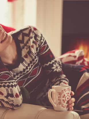 Joyful young woman in a cozy Christmas sweater, sitting in her living room by the fireplace and enjoying a cup of tea on a cold winter's day. She is smiling and looking at camera.