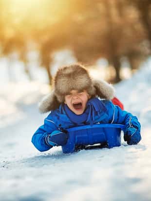 Little boy in funny fake fur hat sliding fast on his sled. The boy aged 6 is screaming with joy. Sunny winter day evening.